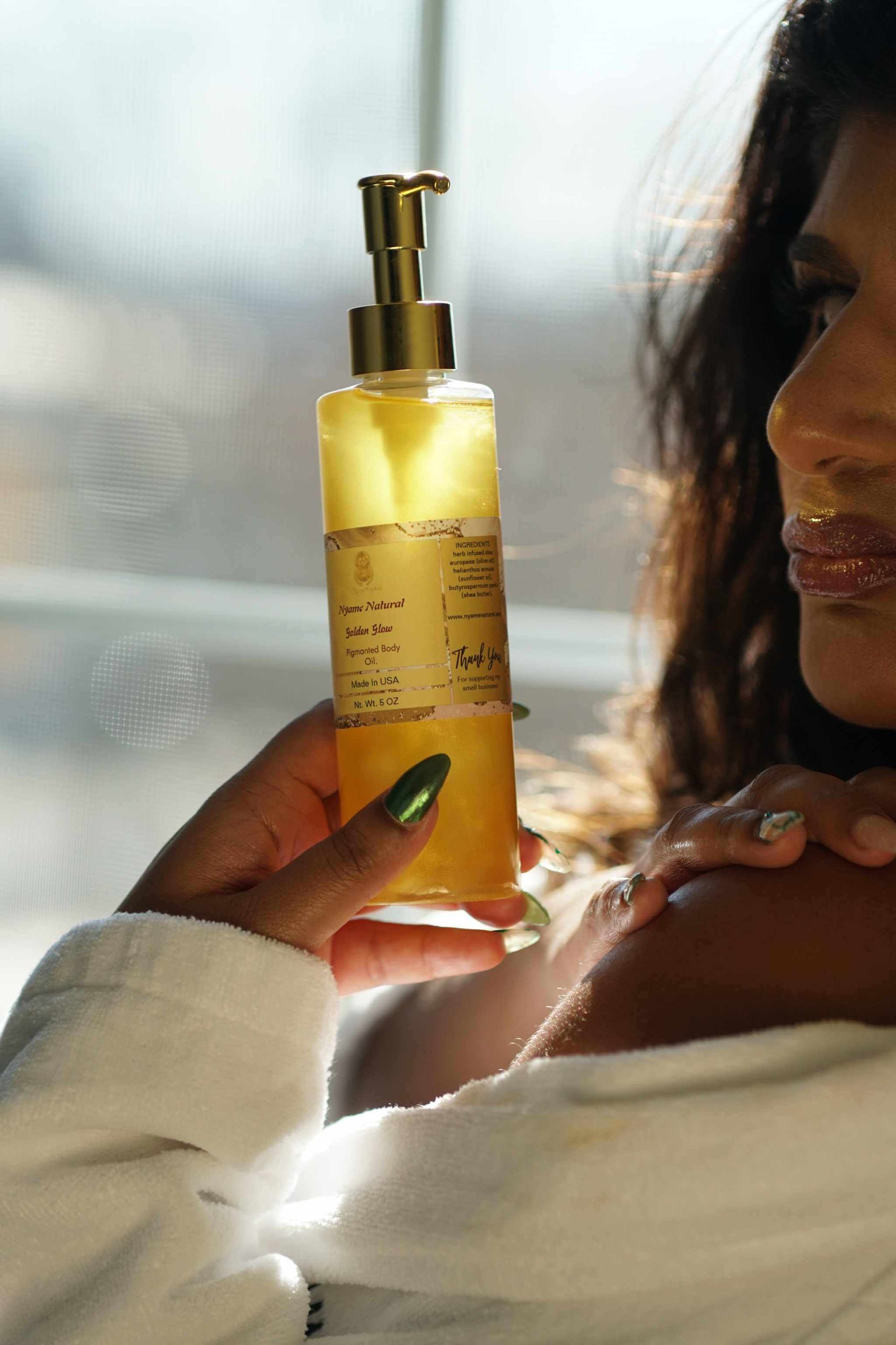 24k Gold Pigmented Pheromone Body Oil in a bottle held by a woman in a robe, showcasing glowing skin and luxurious appeal.