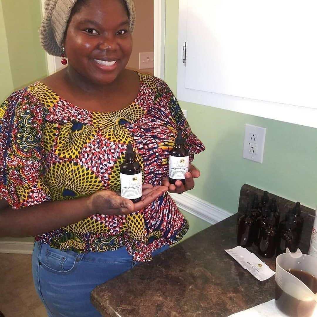 Smiling woman showcasing Haitian Black Castor Oil bottles on kitchen counter, promoting hair growth and healthy hair.