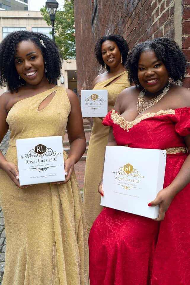 Three women in elegant dresses holding Royal Lux LLC boxes outdoors, showcasing diverse styles and beauty.