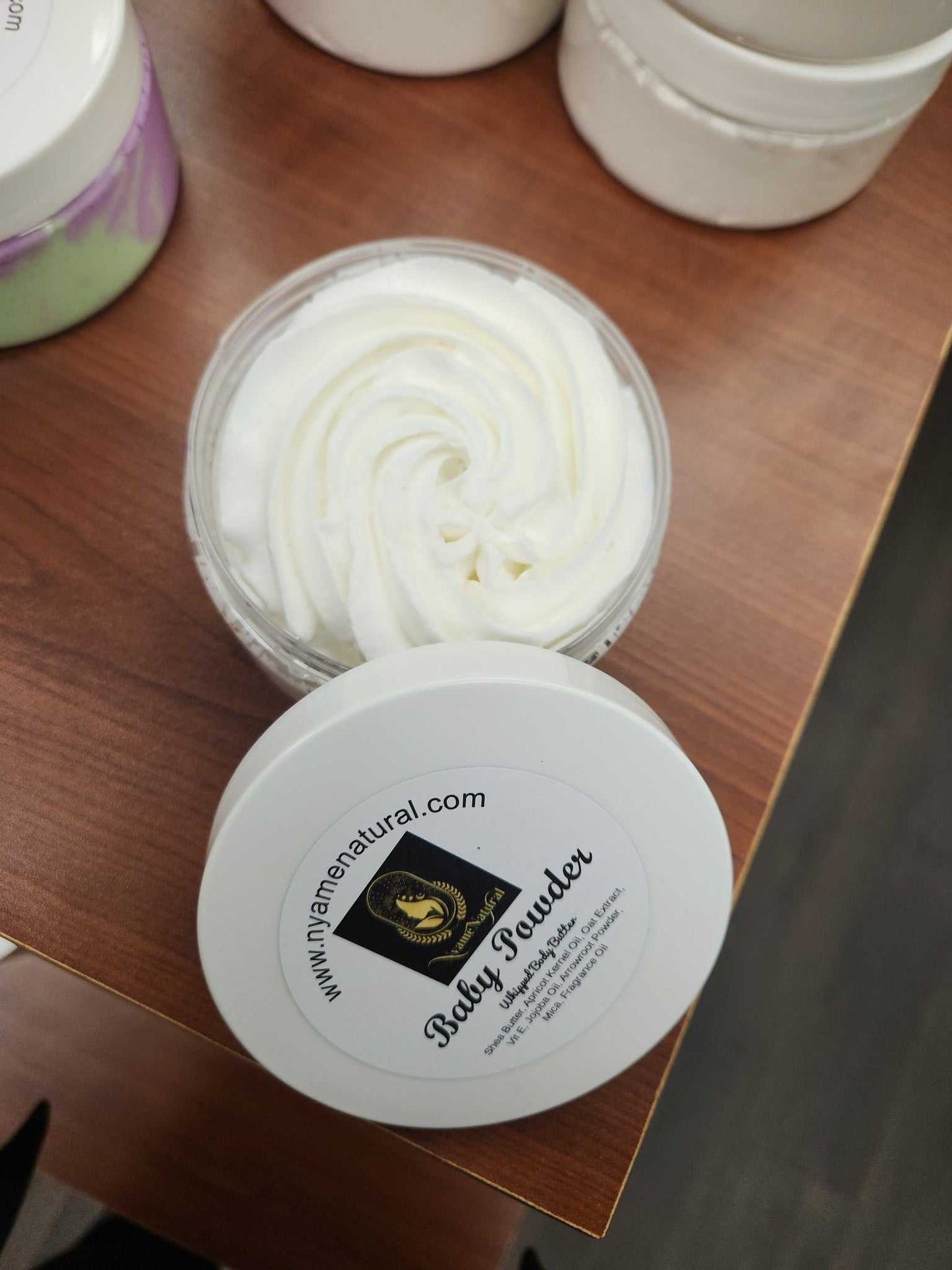 Baby Powder Body Butter in a clear jar with a swirling cream texture on a wooden surface.